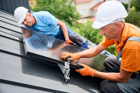 Workers building photovoltaic solar panel system on rooftop of house. Men technicians in helmets and gloves installing solar module with help of hex key outdoors. Alternative energy generation conceptの写真素材