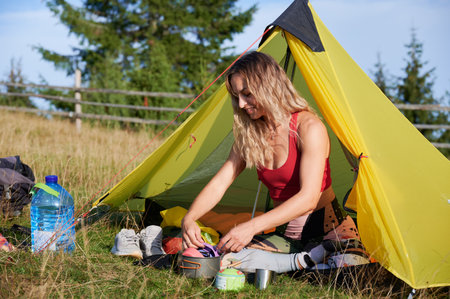 Woman tourist camping outdoors. Female hiker sitting in campsite near tent, cooking, smiling, having break, holding, putting pots. Concept of tourism and hiking in mountains.の写真素材