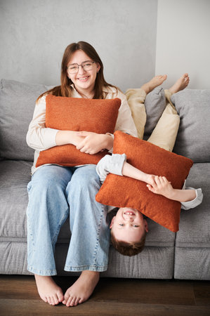 Young siblings relaxing on a couch. Happy sister teenage girl with glasses and brother laying upside down on couch. Smiling kids hugging orange pillows. Adorable children relaxing after pillow fight.の写真素材