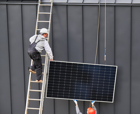 Men installers installing solar panel system on roof of house. Workers in helmets lifting up photovoltaic solar module with help of ropes outdoors. Concept of alternative and renewable energy.の写真素材