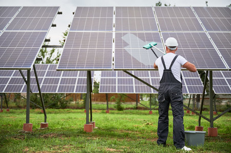 Worker cleaning solar photovoltaic panels by mop. Back view of man taking care about PV battery. Alternative energy innovations concept.の写真素材