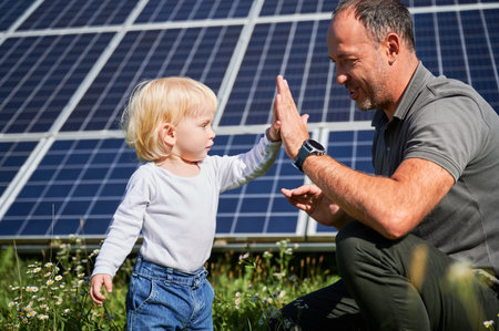 Father and his little son giving each other high-five on background of solar panels. Young father glad spending time with his child. Happy family of two on background of solar station.の写真素材