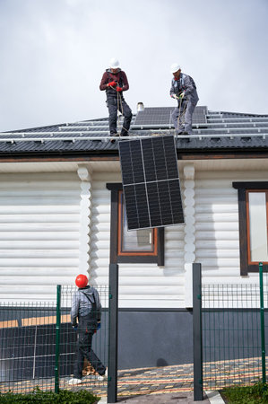 Men workers installing solar panel system on roof of house. Electricians in helmets lifting up photovoltaic solar module with help of ropes outdoors. Concept of alternative and renewable energy.の写真素材