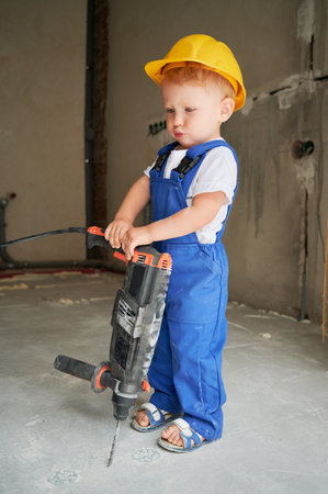 Cute baby boy with electric hammer drill standing in empty room under renovation. Kid construction worker wearing safety helmet and work overalls while holding drilling tool demolition breaker.の写真素材