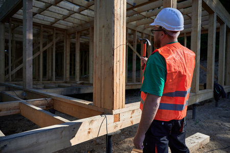 Carpenter constructing two-story wooden frame house near the forest. Back view of man hammering nails into structure while wearing protective helmet and construction vest.の写真素材