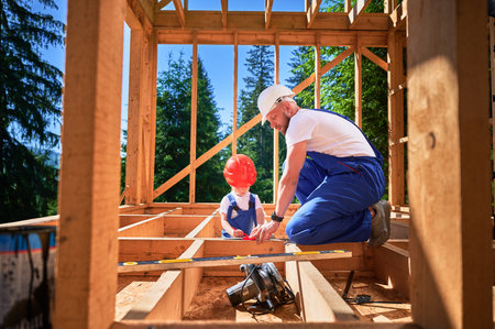 Father with toddler son building wooden frame house. Male builder playing with kid on construction site, wearing helmets and blue overalls on sunny day. Carpentry and family concept.の写真素材
