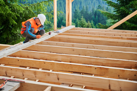 Carpenter in the process of building wooden frame house. Man measures distances with a measuring tape while dressed in workwear and helmet. Concept of modern, environmentally-friendly construction.の写真素材