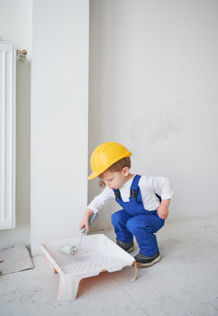 Little boy construction worker crouching down and preparing paint roller for wall painting. Kid wearing safety helmet and work overalls while working on home renovation.の写真素材