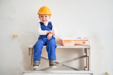 Cute kid construction worker sitting on wooden table against white wall in apartment under renovation. Cheerful little boy wearing safety helmet and work overalls while looking at camera and laughing.の写真素材