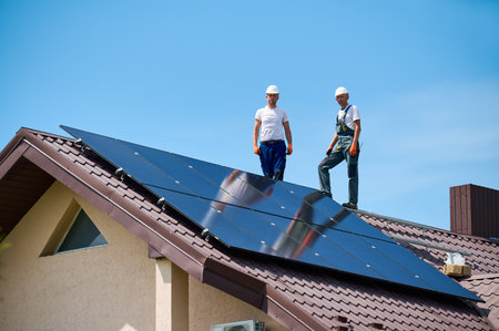 Workers building solar panel system on metal rooftop of house. Portrait of engineers standing on rooftop while installing photovoltaic solar module outdoors. Renewable energy generation concept.の写真素材