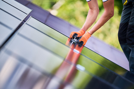 Worker building photovoltaic solar panel system on rooftop of house. Close up of man engineer in gloves installing solar module with help of hex key outdoors. Renewable energy generation concept.の写真素材