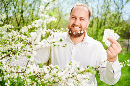 Man allergic suffering from seasonal allergy at spring. Smiling man with small white flowers dotted in beard, holding pack of pills, posing in blossoming garden. Antihistamine medication conceptの写真素材