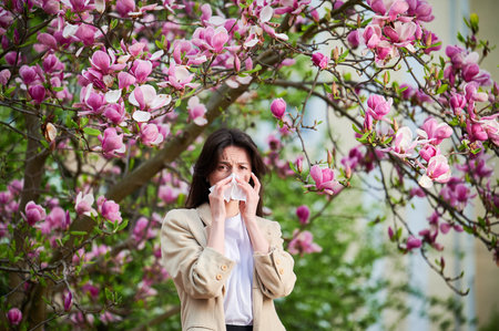 Woman allergic suffering from seasonal allergy at spring in blossoming garden at springtime. Young woman sneezing and blowing nose with nasal handkerchief in front of blooming tree. Allergy concept.の写真素材