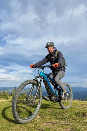 Cyclist man riding electric mountain bike outdoors. Male tourist biking along grassy trail in the mountains, wearing helmet and backpack. Concept of sport, active leisure and nature.の写真素材
