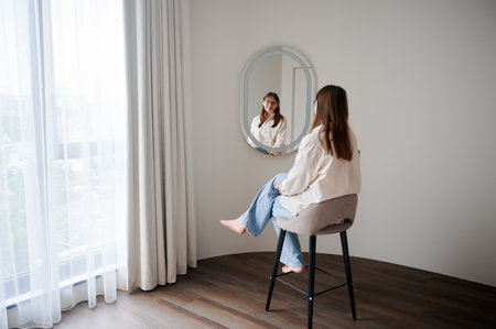 Beautiful girl looking at herself in mirror and smiling. Pretty female checking herself. Cute happy girl sitting in chair opposite to mirror at home.の写真素材