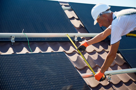 Worker building solar panel system on rooftop of house for generating electricity through photovoltaic effect. Man using ruler to measure mounting equipment for precise installation.の写真素材