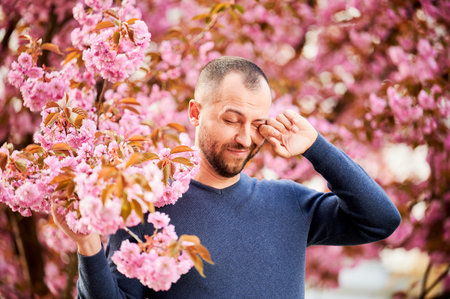 Man allergic suffering from seasonal allergy at spring in blossoming garden at springtime. Bearded man feeling itchy eyes in front of blooming tree. Spring allergy concept.の写真素材
