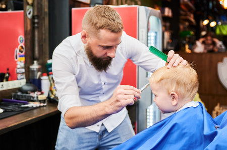 Man barber cutting little boys hair using comb and scissors. Child getting haircut from adult male, likely barber. Professional hairdresser and cute client at modern barbershop.の写真素材