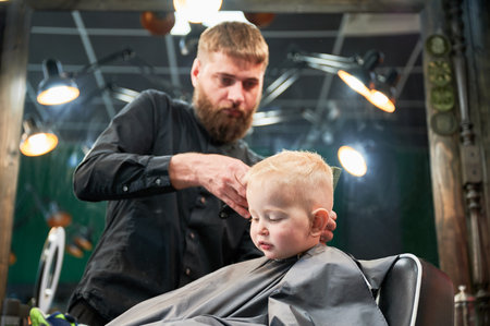 Cute little boy getting haircut. Blond curious kid first time at barbershop. Professional hairdresser making stylish haircut by shaver.の写真素材