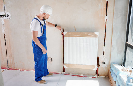 Male plumber in work overalls using wrench tool while installing heating system in apartment under renovation. Man in safety helmet mounting household heating radiator on the wall.の写真素材