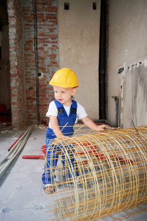 Full length of adorable child in safety helmet and work overalls holding mesh fiberglass rebar. Cute...