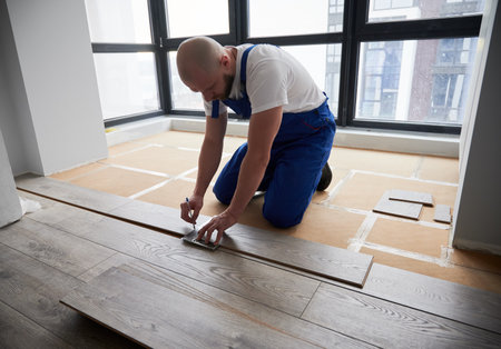 Man in work overalls using metal construction ruler and pen while drawing line on laminate flooring board. Male floor installer preparing laminate plank for installation in apartment under renovation.の写真素材