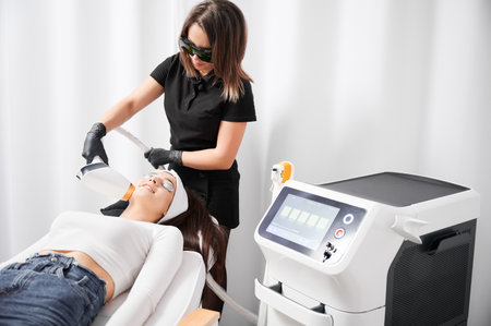 Female cosmetologist in protective glasses using diode laser device while performing skin rejuvenation procedure in clinic. Woman lying near cosmetology equipment during beauty procedure.の写真素材