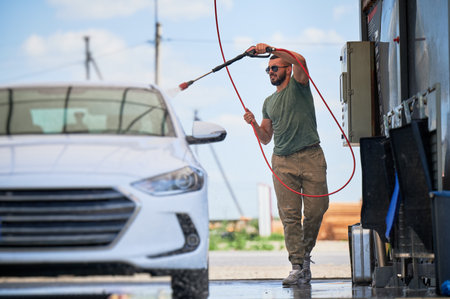Washing luxury white auto with water gun on an open air car wash. Confident man cleaning his car with high pressure water jet. Technical service concept.の写真素材