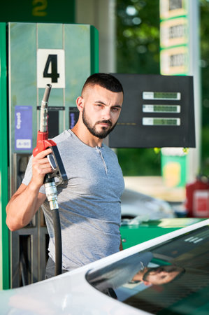 Skilled driver refueling his automobile for long road. Portrait of male adult with pump nozzle refueling car tank. Man holding gasoline pump next to car on background gas column.の写真素材
