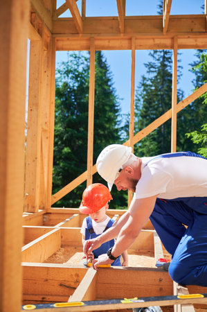 Father with toddler son building wooden frame house. Male builder playing with kid on construction site, wearing helmets and blue overalls on sunny day. Carpentry and family concept.の写真素材