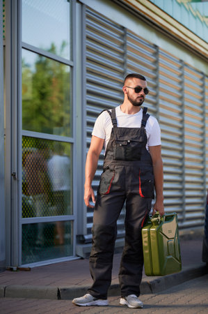 Strong gas station worker holding canister full of fuel. Male operator caring heavy cistern with gasoline. Confident man in sunglasses and overalls with canister looking away.の写真素材