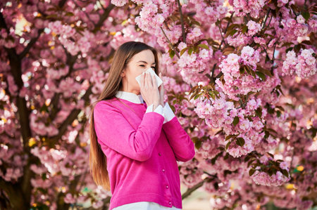 Woman allergic suffering from seasonal allergy at spring in blossoming garden at springtime. Woman sneezing and blowing nose using nasal handkerchief in front of blooming tree. Spring allergy concept.の写真素材