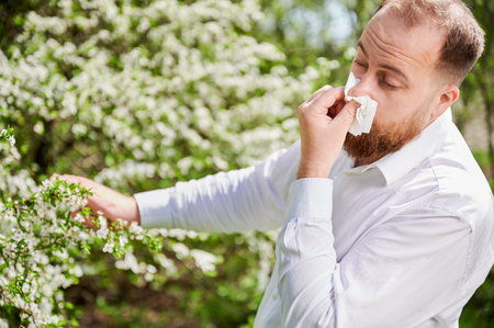 Man allergic suffering from seasonal allergy at spring in blossoming garden at springtime. Young man sneezing and blowing nose with nasal handkerchief in front of blooming tree. Spring allergy conceptの写真素材