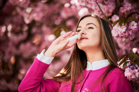 Woman allergic using medical nasal drops, suffering from seasonal allergy at spring in blossoming garden. Close up of woman treating runny nose near blooming tree outdoors. Spring allergy concept.の写真素材