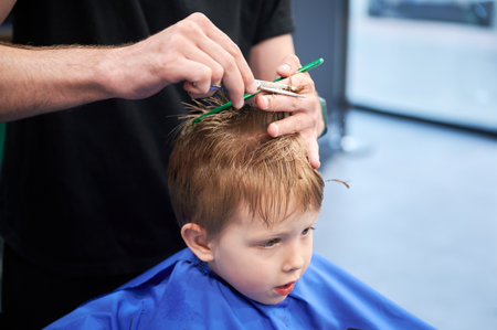 Man barber cutting little boys hair using comb and scissors. Child getting haircut from adult male, likely barber. Professional hairdresser and cute client at modern barbershop.の写真素材