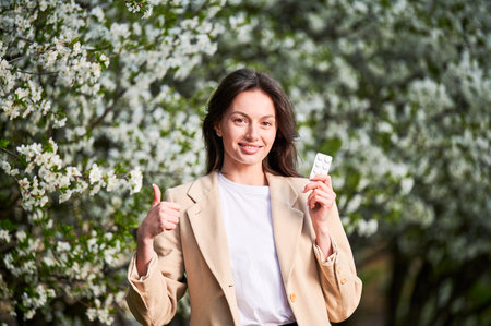 Woman allergic suffering from seasonal allergy at spring. Young happy woman showing allergy drugs, giving thumbs up, posing in blossoming garden at springtime. Antihistamine medication conceptの写真素材
