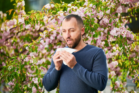 Man allergic suffering from seasonal allergy at spring in blossoming garden at springtime. Man sneezing and blowing nose using nasal handkerchief in front of blooming tree. Spring allergy concept.の写真素材