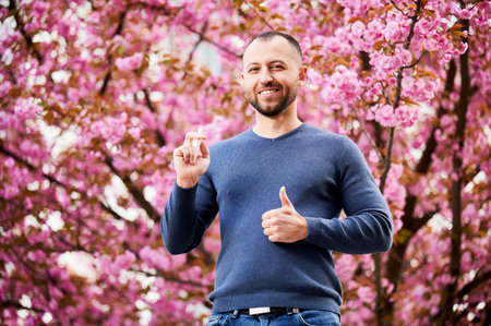 Man allergic using medical nasal drops, suffering from seasonal allergy at spring in blossoming garden. Portrait of smiling man showing thumbs up near blooming tree outdoors. Spring allergy concept.の写真素材