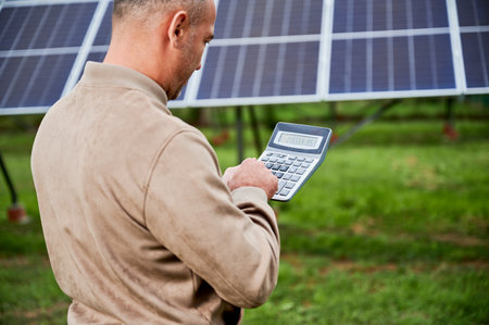 Professional investor calculating his possible profit from alternative enegry sources. Man using calculator to count exact sum of income. Back view of man counting on background of solar panels.の写真素材