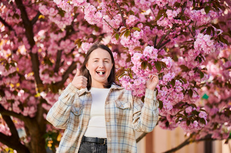 Woman allergic suffering from seasonal allergy at spring. Young happy woman applying allergy pill, giving thumbs up, posing in blossoming garden at springtime. Antihistamine medication conceptの写真素材