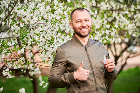 Man allergic using medical eyes drops, suffering from seasonal allergy at spring in blossoming garden. Portrait of smiling man showing thumbs up in front of blooming tree. Spring allergy concept.の写真素材