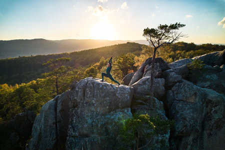 Woman practicing yoga and meditation on the top of huge boulder in the evening. Beautiful sunset, autumn forests, rocks and hills on background. Aerial drone viewの写真素材