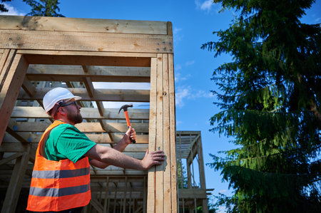 Carpenter constructing wooden frame house near the forest. Bearded man hammering nails into structure while wearing protective helmet and construction vest. Concept of modern ecological construction.の写真素材