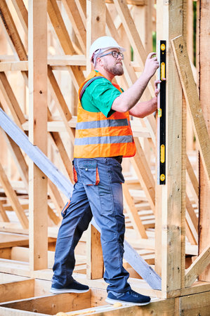 Carpenter constructing two-storey wooden frame house. Man inspects walls for levelness using spirit level, wearing protective overalls, helmet and vest. Concept of modern ecological construction.の写真素材