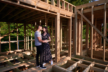Man and woman appraising their future wooden frame dwelling nestled near the forest. Youthful couple at construction site in early morning. Concept of contemporary ecological construction.の写真素材