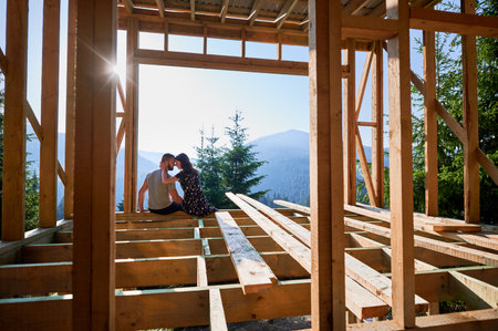 Man and woman inspecting their future wooden frame dwelling nestled in the mountains near forest. Youthful couple at construction site in the morning. Concept of contemporary ecological construction.の写真素材