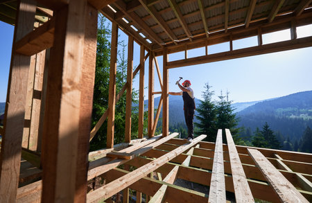 Carpenter constructing wooden frame, two-story house near forest. Man hammering nails with hammer while dressed in protective helmet, work coveralls. Concept of eco-friendly modern construction.の写真素材