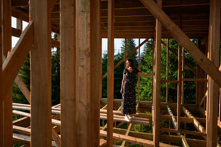 Woman inspecting future wooden frame dwelling nestled in the mountains near forest. Youthful female at construction site in early morning. Concept of contemporary ecological construction.の写真素材