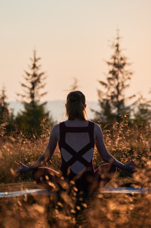 Back view of sporty girl sitting in lotus pose. Young female doing yoga with view on mountains. Meditation at sunrise.の写真素材