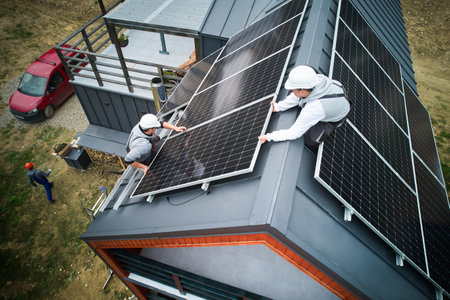 Roofers building photovoltaic solar module station on roof of house. Men electricians in helmets installing solar panel system outdoors. Concept of alternative and renewable energy. Aerial view.の写真素材